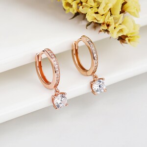 May include: Rose gold hoop earrings with a dangling crystal accent. The hoop earrings are adorned with small clear stones.