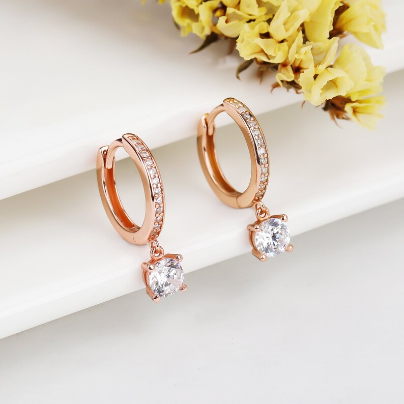 May include: Rose gold hoop earrings with a dangling crystal accent. The hoop earrings are adorned with small clear stones.