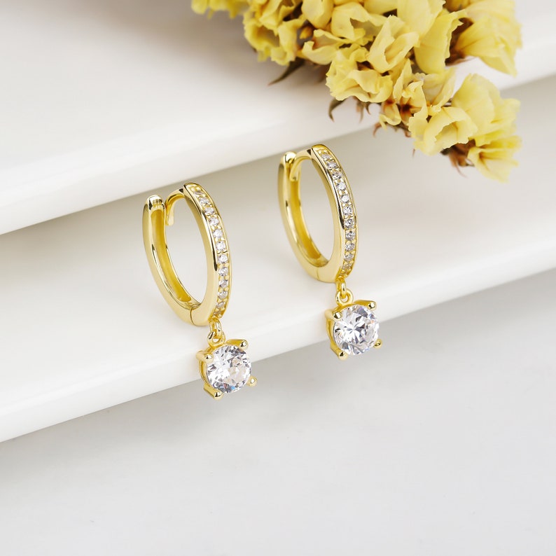 May include: A pair of gold hoop earrings with a dangling crystal. The hoops are lined with small clear stones.
