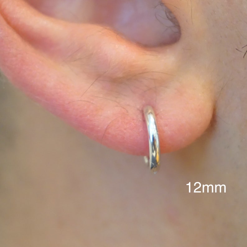 May include: A small, polished silver-colored hoop earring inserted into an earlobe. The hoop has a smooth, rounded surface and a simple circular design. The text 12mm is clearly visible in the lower right corner of the image.
