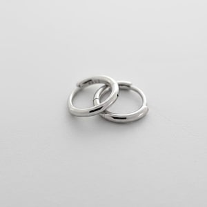 May include: A pair of small, polished silver-tone hoop earrings with a smooth, rounded profile. The earrings are interlocking, featuring a hinged closure mechanism that is visible on the side of the circular metal bands.