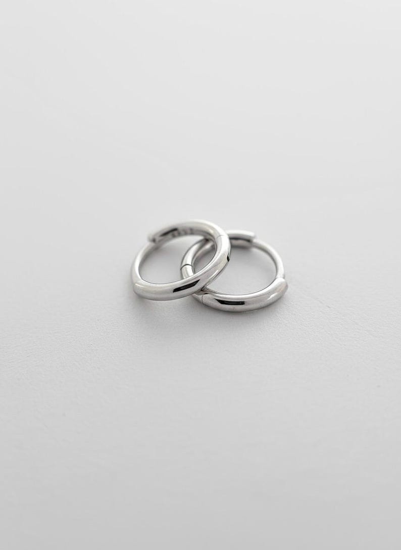 May include: A pair of small, polished silver-tone hoop earrings with a smooth, rounded profile. The earrings are interlocking, featuring a hinged closure mechanism that is visible on the side of the circular metal bands.