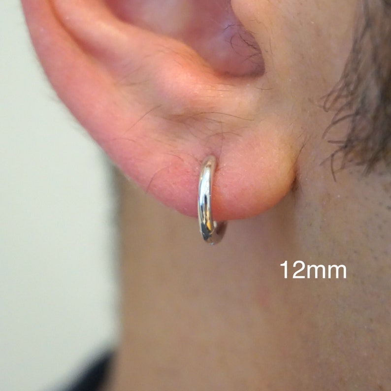 May include: A small, polished silver hoop earring worn in an earlobe. The earring has a smooth, circular design with a reflective metallic finish. The text 12mm is clearly visible in the lower right corner, indicating the size of the hoop.