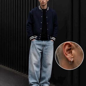 May include: A navy blue varsity jacket with white striped ribbed cuffs and collar, worn over a black shirt and light blue baggy denim jeans. A small circular inset shows a silver hoop earring worn in the earlobe of the individual.