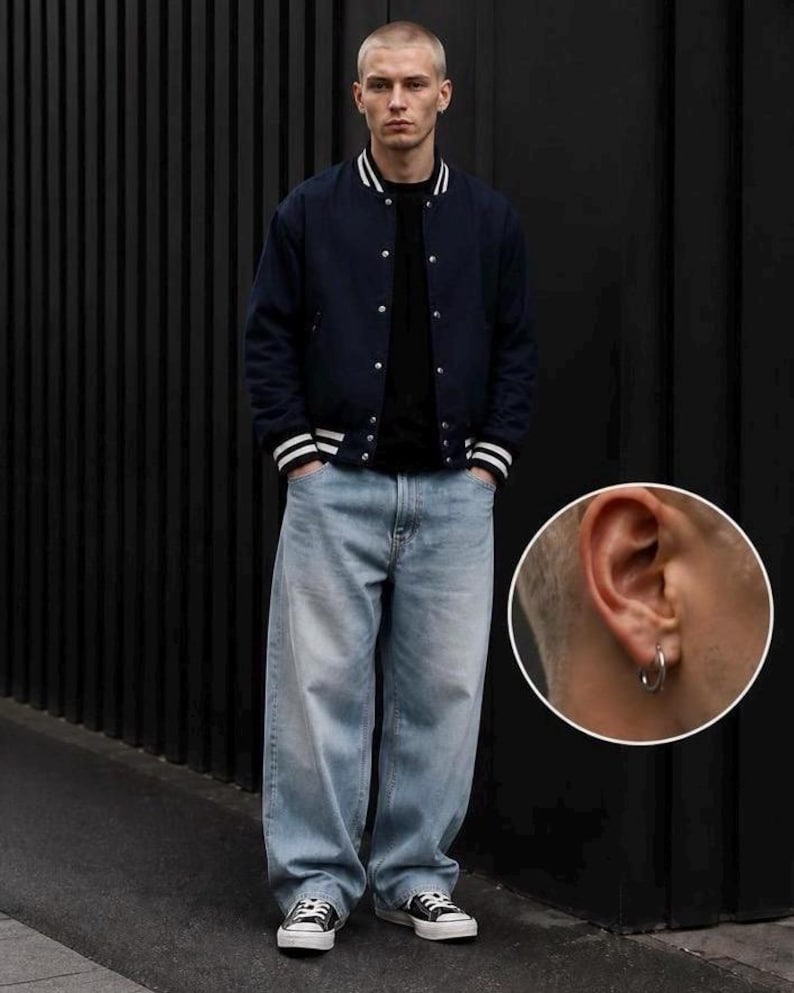 May include: A navy blue varsity jacket with white striped ribbed cuffs and collar, worn over a black shirt and light blue baggy denim jeans. A small circular inset shows a silver hoop earring worn in the earlobe of the individual.