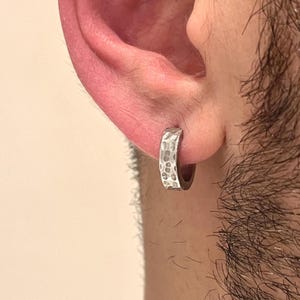 Pode incluir: Um pequeno brinco de argola circular com acabamento em tom prateado e uma superfície texturizada e martelada. O brinco apresenta um design de banda grossa que se ajusta ao lóbulo, exibindo um padrão metálico irregular e reflexivo.