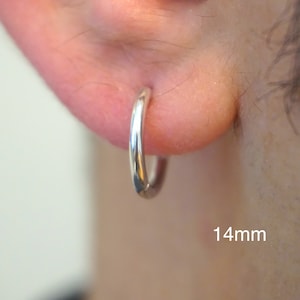May include: A polished silver hoop earring with a smooth, circular design. The earring is shown inserted into an earlobe, with the text 14mm clearly visible in the lower right corner, indicating the diameter of the hoop earring.