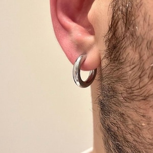 May include: A polished silver-tone hoop earring with a thick, rounded profile. The circular earring features a smooth, reflective metallic surface and is worn through the earlobe, showcasing a simple and clean design without any additional embellishments.