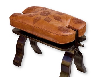 May include: A brown leather stool with a unique shape, featuring a carved floral pattern on the seat. The stool is supported by dark wooden legs and a lower shelf. The seat measures approximately 24 inches long.