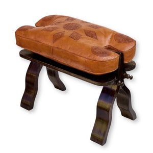 Pode incluir: Um banco de couro castanho com uma forma única, apresentando um padrão floral esculpido no assento. O banco é suportado por pernas de madeira escura e uma prateleira inferior. O assento mede aproximadamente 61 cm de comprimento.