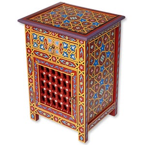 Peut inclure: Une table de chevet en bois peinte à la main avec des motifs géométriques complexes en rouge, or, bleu et violet. Elle comporte un tiroir et une porte avec un motif en treillis. C'est un meuble décoratif.
