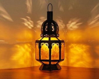 May include: A black metal lantern with a glass panel and a warm yellow glow. The lantern has a decorative design and is sitting on a wooden surface.