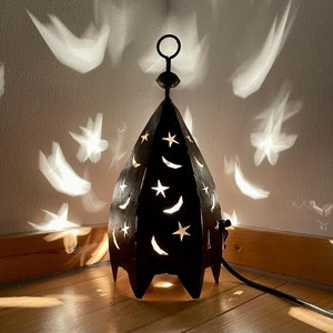 May include: A black metal lamp with a star and crescent moon design. The lamp is lit and casting shadows on the wall.