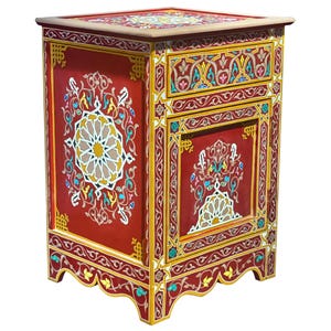 Op de afbeelding: Een rood, handbeschilderd houten nachtkastje met ingewikkelde bloemen- en geometrische ontwerpen in geel, wit en blauw. Het nachtkastje heeft een enkele lade en een decoratief bovenblad.