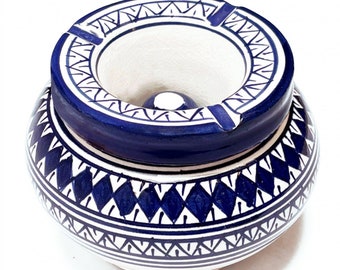 May include: A white ceramic ashtray with a blue and white geometric pattern. The ashtray has a round top with a small indentation in the center and a wider base.