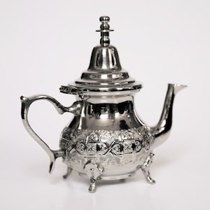 Könnte beinhalten: Eine silberne Teekanne mit einem verzierten Design und einem Deckel. Die Teekanne hat einen Ausguss und einen Griff. Sie hat drei Beine und ist aus Metall.