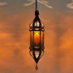 Può includere: Una lanterna sospesa ornata, con una struttura in metallo scuro e intricati motivi traforati. L'interno della lanterna emette una calda luce ambrata, creando un'atmosfera accogliente. Ideale per l'arredamento della casa.