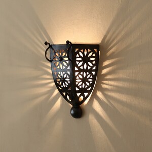 K&ouml;nnte beinhalten: Eine schwarze Wandleuchte aus Metall mit geometrischem Design. Die Leuchte hat eine Gl&uuml;hbirne im Inneren und wirft einen Schatten an die Wand.
