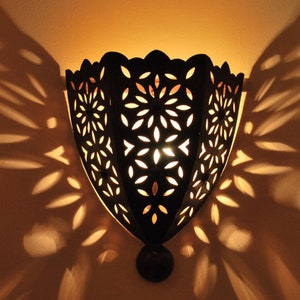 Op de afbeelding: Een zwarte metalen wandlamp met een uitgesneden ontwerp van herhalende bloemenvormen. De lamp is verlicht en werpt ingewikkelde schaduwen op de muur.