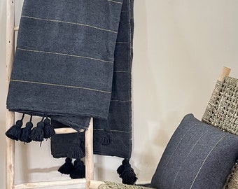May include: A dark gray throw blanket with gold stripes and black tassels, draped over a wooden ladder. A matching pillow rests on a woven chair, creating a cozy, neutral-toned aesthetic.