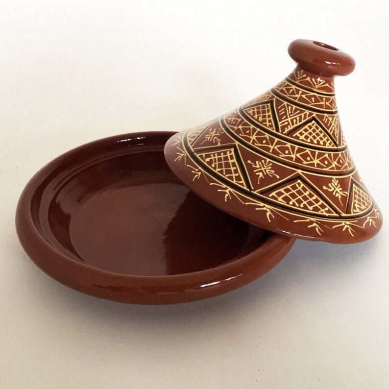 May include: A brown ceramic tagine with a lid featuring a geometric pattern in yellow and black. The tagine is a traditional Moroccan cooking pot.