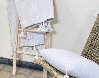May include: A white throw blanket with white pom-poms on a wooden ladder. A white pillow with white pom-poms is on a woven chair.