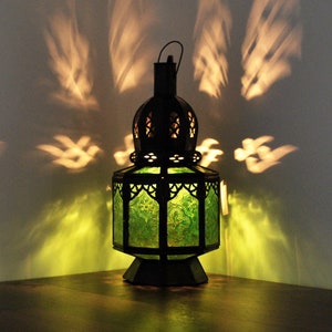Può includere: Una lanterna decorativa in metallo nero con pannelli in vetro verde. La lanterna ha un manico e intricati disegni traforati, che proiettano luce a motivi sul muro. La lanterna è posizionata su una superficie di legno, creando un'atmosfera calda.