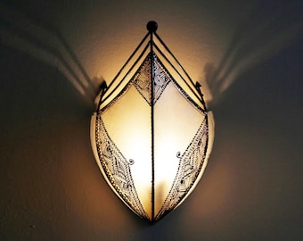 May include: A decorative wall sconce with a cream-colored, shield-shaped shade. The shade is framed by black metal and features intricate black patterns. The sconce is illuminated, casting shadows on the wall. The fixture is designed for indoor use.