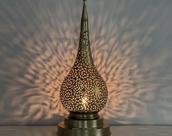 May include: A decorative brass table lamp with a pierced, teardrop-shaped shade. The lamp's intricate cut-out design casts a pattern of light on the wall. The lamp has a round base and a tall, pointed top. The website address www.L-artisan.de is visible on the base.