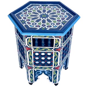 Può includere: Un tavolino esagonale fatto a mano con un design geometrico vibrante. Il tavolo presenta un piano blu scuro con un motivo a stella in bianco, verde e rosso. I lati sono intagliati e dipinti con motivi simili, creando un mobile d'arredo.