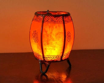 May include: An orange barrel-shaped lamp with black tribal designs and three small legs. The lamp emits a warm, inviting glow. The design includes geometric patterns and floral motifs, creating a unique decorative piece.