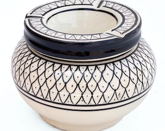 May include: Cream-colored ceramic ashtray with a black and white geometric design. The ashtray has a round shape with a wide rim and a central bowl. The exterior features a repeating pattern of black teardrop shapes. The rim is black with a white geometric border.