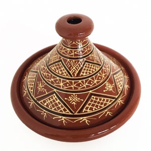 May include: A terracotta tagine with a conical lid featuring intricate geometric patterns in gold and black. The tagine is a traditional Moroccan cooking pot.