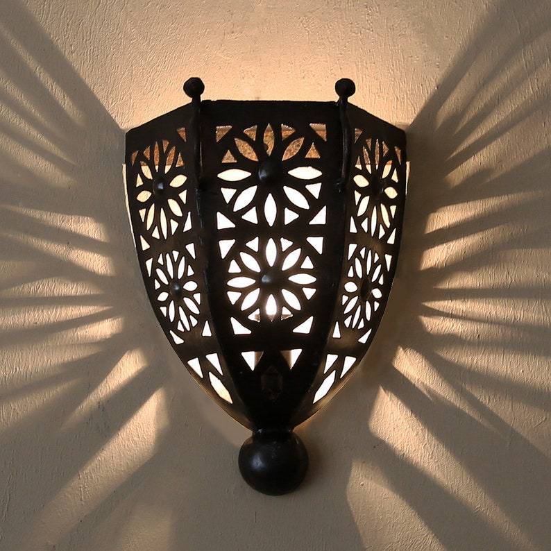 K&ouml;nnte beinhalten: Eine schwarze Wandleuchte aus Metall mit geometrischem Design und einer Gl&uuml;hbirne im Inneren. Die Leuchte wirft komplizierte Schatten an die Wand.