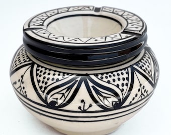 May include: A ceramic ashtray with a cream-colored base and black geometric patterns. The ashtray has a circular design with a recessed area for holding ashes and cigarettes. The rim and the body are decorated with intricate black designs.