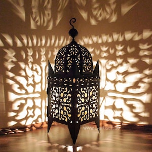 Può includere: Una lanterna in metallo nero con intricati motivi traforati, che proietta ombre decorative sulla parete. La lanterna ha una parte superiore a cupola e una base con piedi a punta. La calda luce all'interno crea un'atmosfera accogliente.