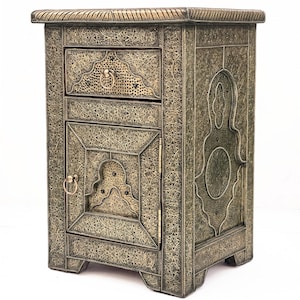 Op de afbeelding: Oud nachtkastje met een lade en een kastdeur. De metalen kast heeft een gedempte goudkleur en is voorzien van ingewikkelde, in reliëf aangebrachte patronen over het oppervlak. De lade en de deur hebben ringtrekkers. De kast is ongeveer 61 cm hoog.