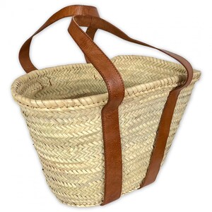 May include: A woven straw tote bag with brown leather handles. The bag is large and has a rectangular shape.