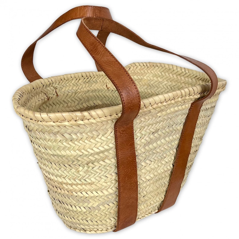 May include: A woven straw tote bag with brown leather handles. The bag is large and has a rectangular shape.