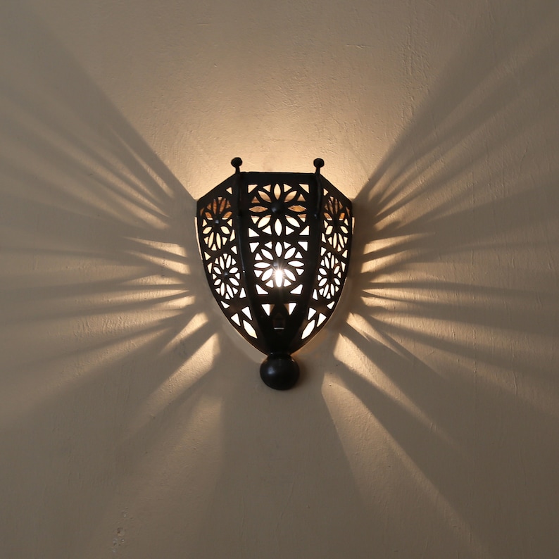 K&ouml;nnte beinhalten: Eine schwarze Wandleuchte aus Metall mit geometrischem Design und einer Gl&uuml;hbirne im Inneren. Die Leuchte wirft einen Schatten an die Wand.