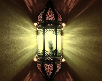 May include: A wall-mounted lantern featuring a dark metal frame with intricate geometric cutouts. The lantern emits a bright, warm yellow light through its panels, casting distinct, elongated shadow patterns onto the surrounding wall surface.