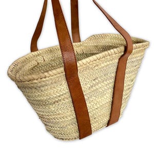 May include: A natural woven straw tote bag with brown leather straps. The bag is oval shaped and has a wide opening.
