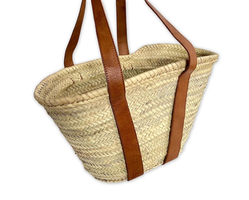 May include: A natural woven straw tote bag with brown leather straps. The bag is oval shaped and has a wide opening.