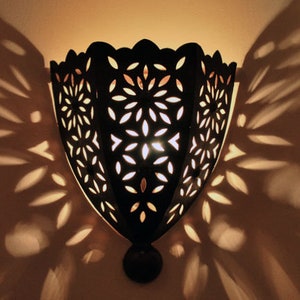 Op de afbeelding: Een zwarte metalen wandlamp met een decoratief uitgesneden ontwerp. De lamp werpt ingewikkelde schaduwen op de muur.
