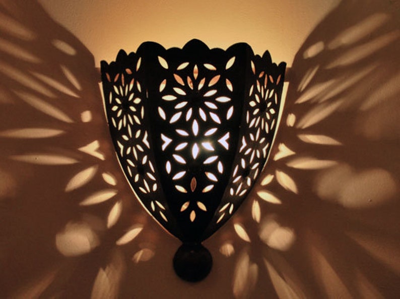 Op de afbeelding: Een zwarte metalen wandlamp met een decoratief uitgesneden ontwerp. De lamp werpt ingewikkelde schaduwen op de muur.