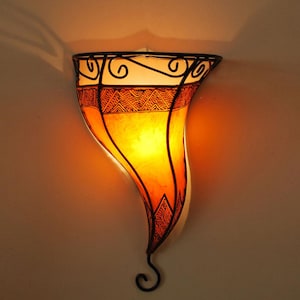 Puede incluir: Una lámpara de pared con una pantalla curva de color naranja y blanco. La pantalla tiene un marco de metal decorativo con diseños intrincados.