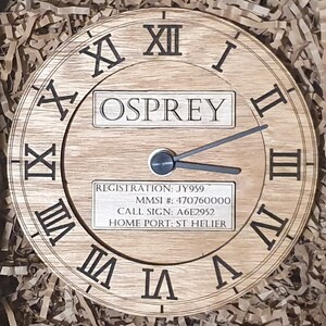 May include: A wooden clock face with Roman numerals and a black clock hand. The clock face has a label that reads "OSPREY" and includes the following information: "REGISTRATION: JY959", "MMSI #: 470760000", "CALL SIGN: A6E2952", "HOME PORT: ST HELIER".