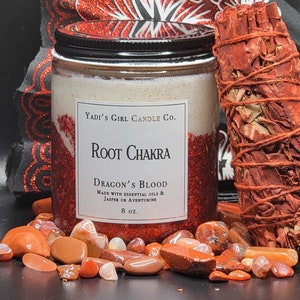 May include: A glass jar candle with a black lid, labeled "Root Chakra Dragon's Blood" with essential oils and jasper or aventurine. The candle is surrounded by red stones and a bundle of dried herbs. The candle is 8 oz.