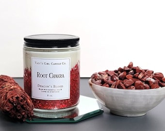 Könnte beinhalten: Eine rote und weiße Kerze in einem Glasgefäß mit schwarzem Deckel. Die Kerze ist mit "Root Chakra" und "Dragon's Blood" beschriftet. Die Kerze steht auf einem silbernen Tablett mit roten Steinen in einer weißen Schale daneben.