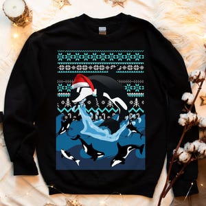 Könnte beinhalten: Schwarzer Weihnachtspullover mit einem blauen und weißen Schneeflockenmuster. Eine schwarz-weiße Orca, die einen Weihnachtsmann-Hut trägt, springt aus dem Wasser, während andere Orcas um sie herum schwimmen.